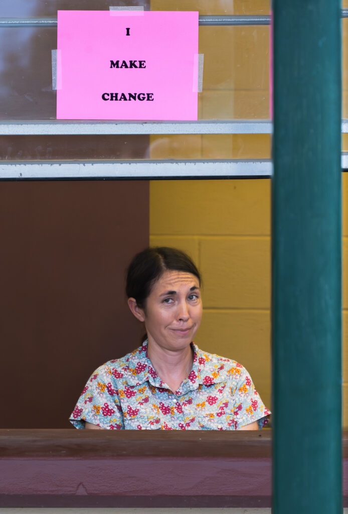 Woman in floral shirt behind counter with pink I MAKE CHANGE sign on yellow brown wall