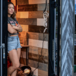 Woman standing in a small wooden-paneled room with an open door, fan, and table inside.