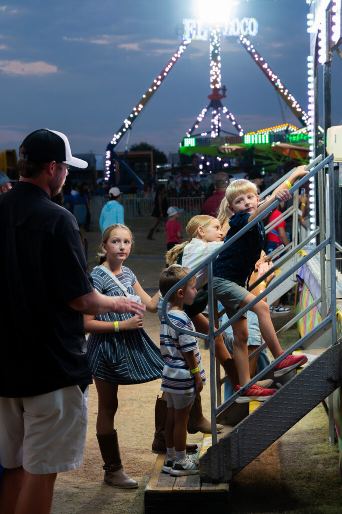 Children and an adult waiting and climbing stairs to a carnival ride at dusk with lit rides in background