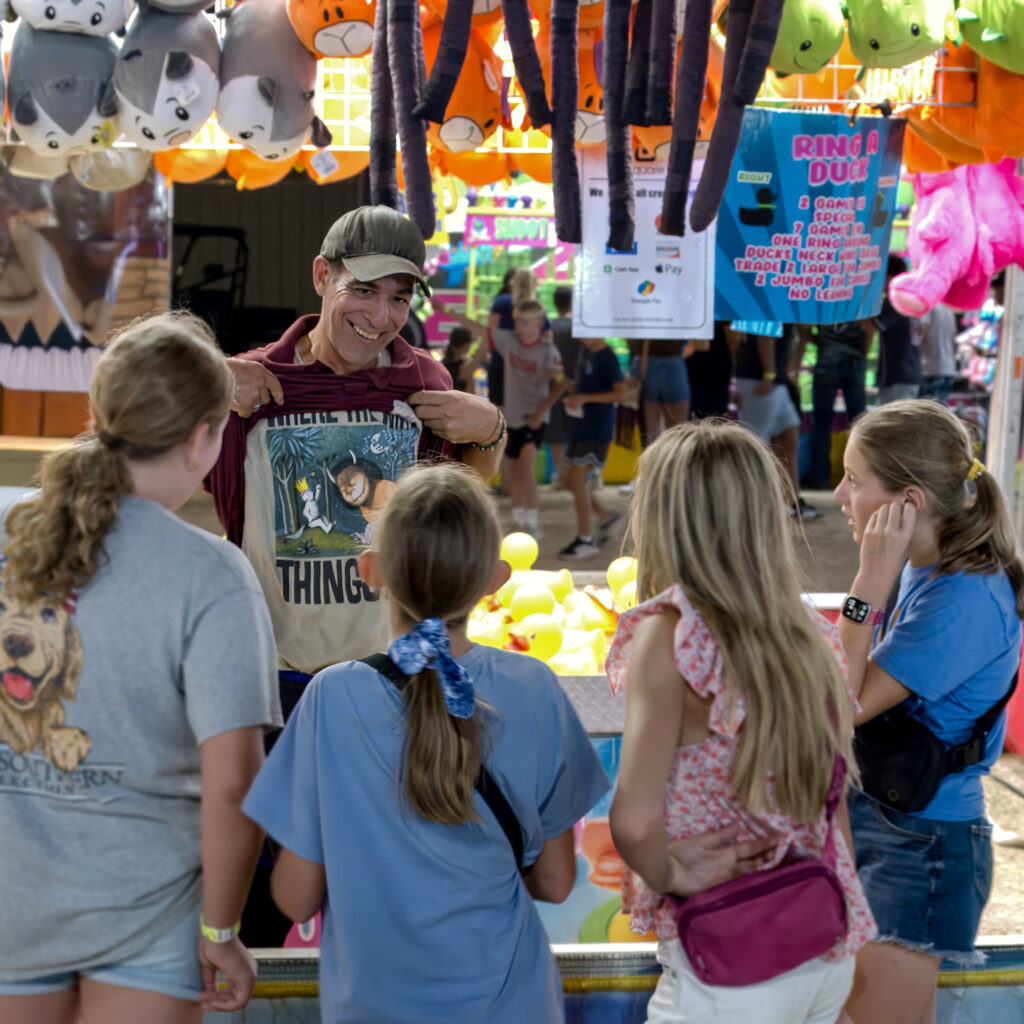 Man at carnival booth showing a printed shirt to four girls near a Ring a Duck game.