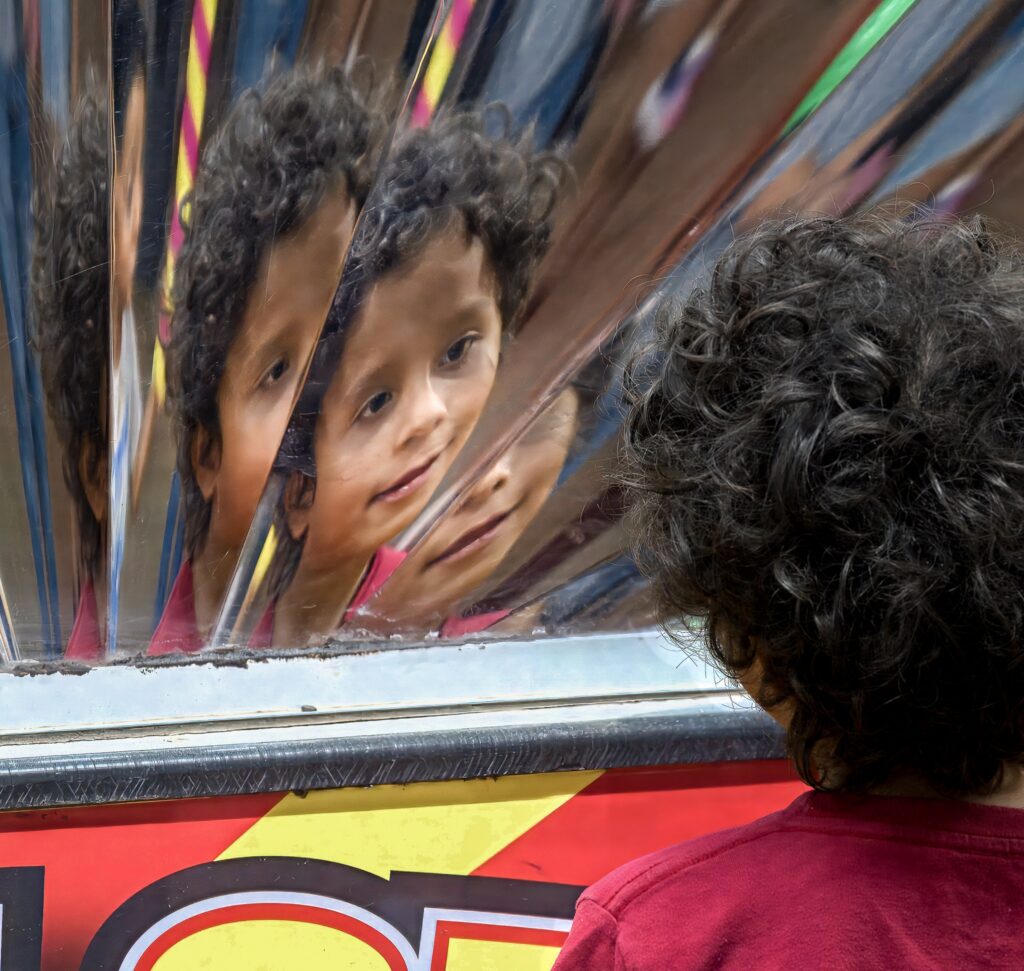 Child with curly hair in a red shirt viewing their distorted reflection in a segmented mirror.