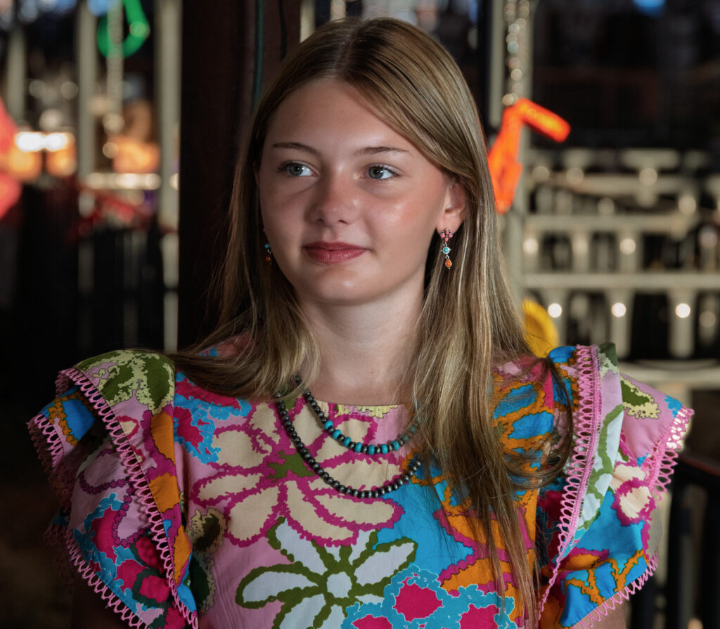 Young woman indoors wearing a colorful floral dress, beaded necklaces, and dangling earrings.