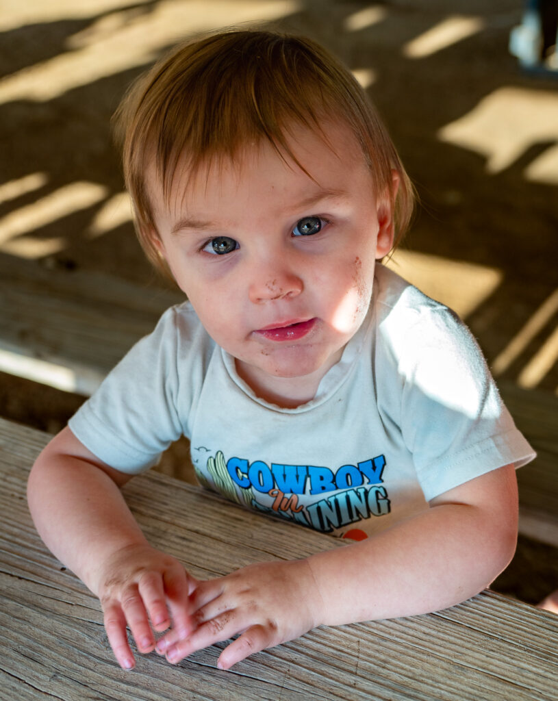 Child with dirt on face and hands wearing Cowboy in Training shirt sitting at wooden table.