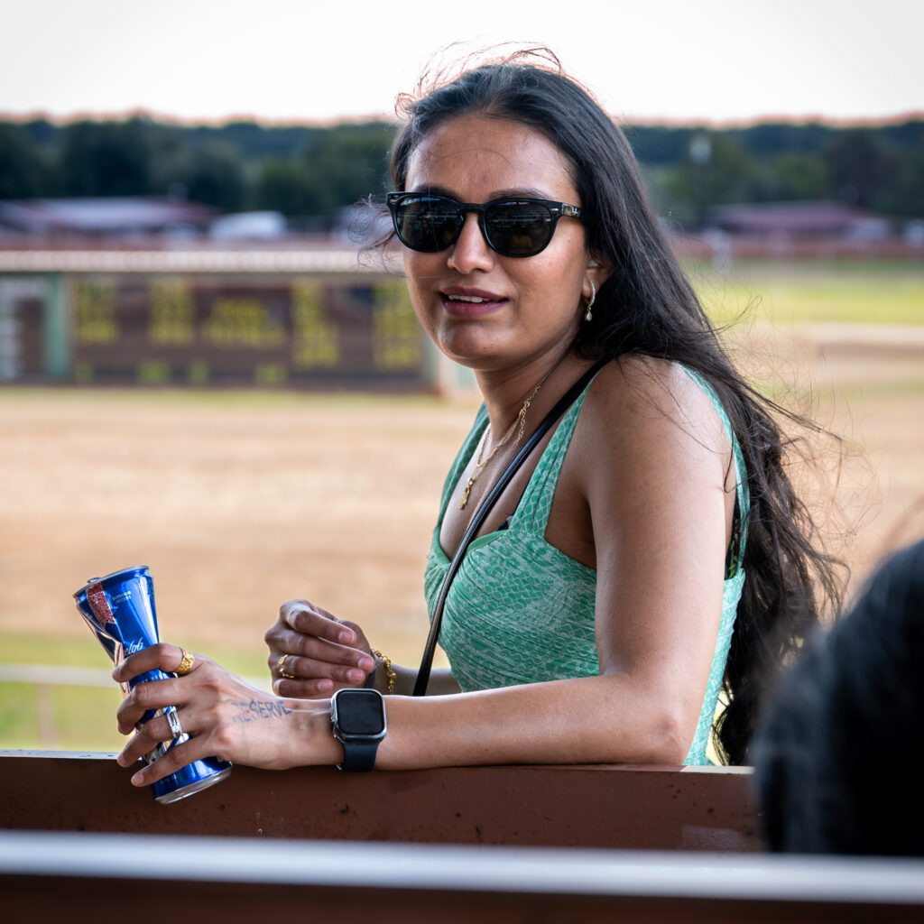 Woman with sunglasses and smartwatch holding blue can outdoors near a railing with a field background