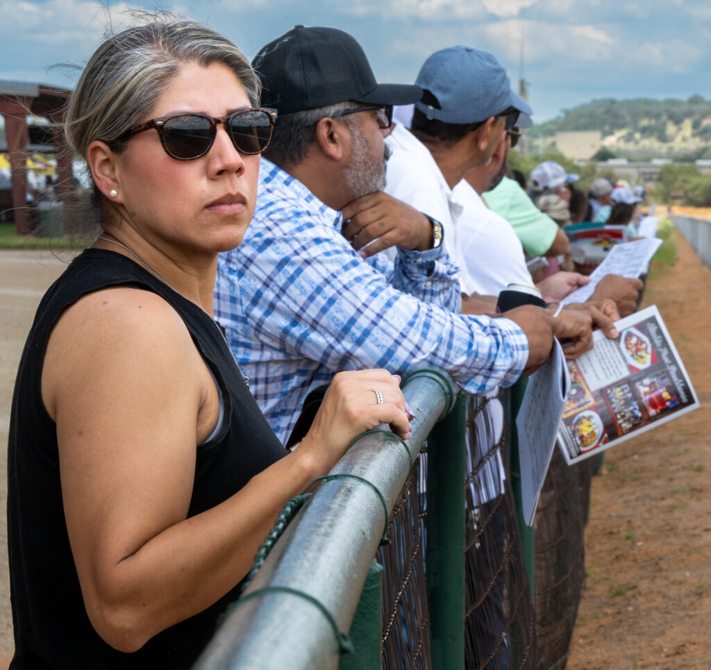 People standing along a fence outdoors, some holding papers, watching an event on a dirt path.