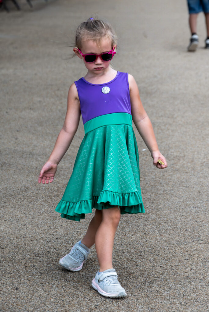 Young girl wearing a purple and green dress with pink sunglasses and gray athletic shoes walking.