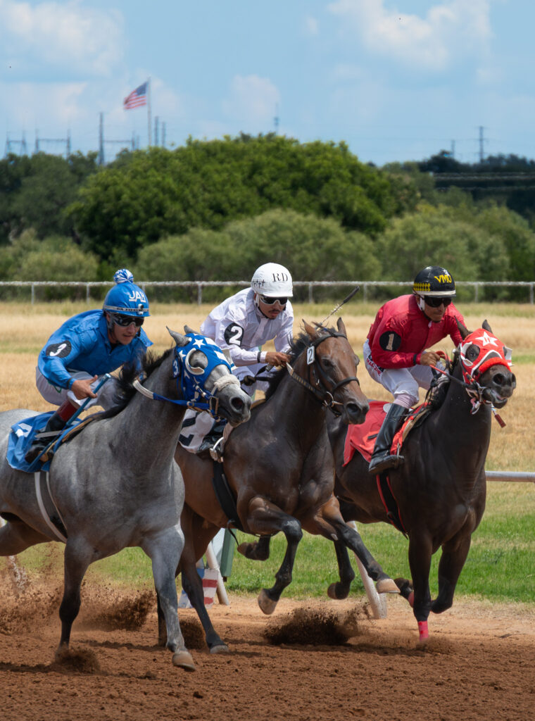 Three jockeys in blue, white, and red silks racing horses closely on a dirt track.