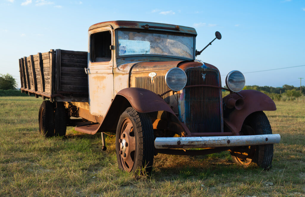 Rusty vintage flatbed truck with wooden cargo bed parked on grassy field under clear sky.