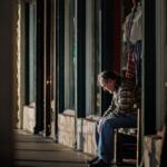 Older man in plaid shirt and jeans sitting on a wooden bench outside a storefront.