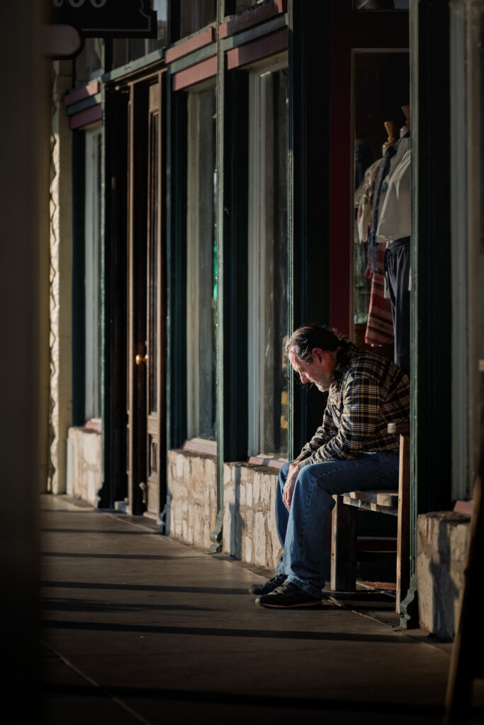 Older man in plaid shirt and jeans sitting on a wooden bench outside a storefront.
